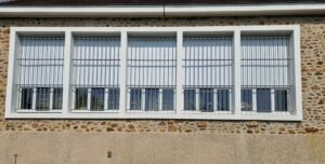 Store extérieur à descente verticale à motorisation électrique - Toile occultante - Armature et toile colorie blanc - Installée sur 5 fenêtres de derrière une grille de protection